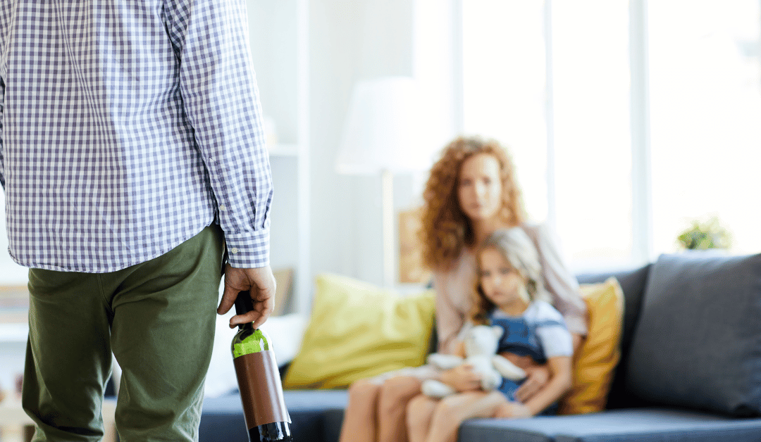 Back view of husband with a half-empty wine bottle in his hand while is worried wife and child sit on the couch in fear.