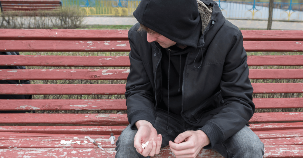 Man in a hoodie sitting on a park bench with needles and drugs next to him.