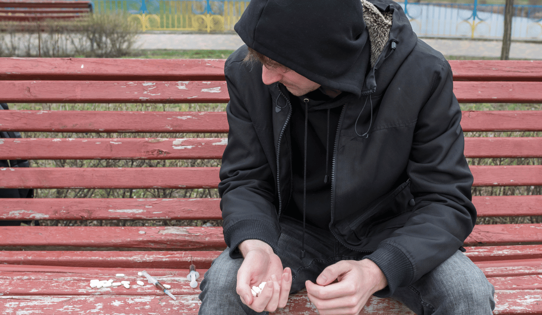 Man in a hoodie sitting on a park bench with needles and drugs next to him.