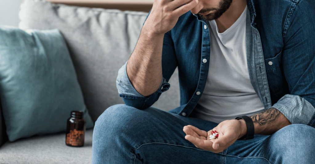 Man sitting on a couch with his head in one of his hands, a handful of pills in the other and a bottle of pills on the couch next to him.