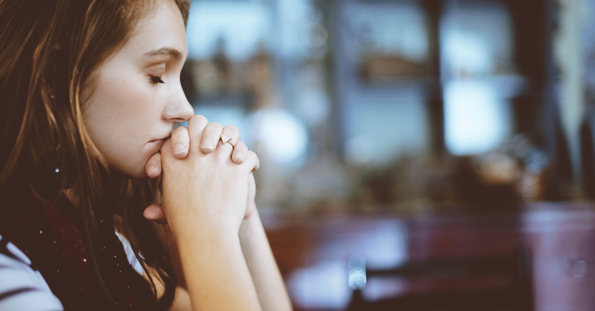 Woman sitting at a table with her elbows on the table and her hands clasped in front of her face and her eyes closed. She appears to be hoping and praying.