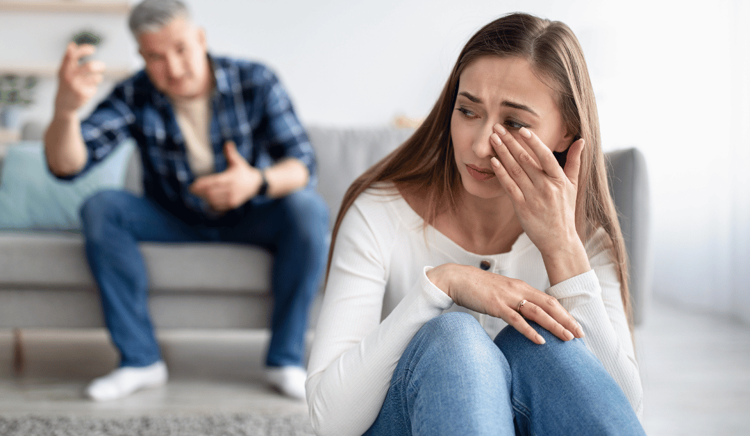 Crying woman sitting on the floor with her husband sitting on the couch behind her yelling at her.